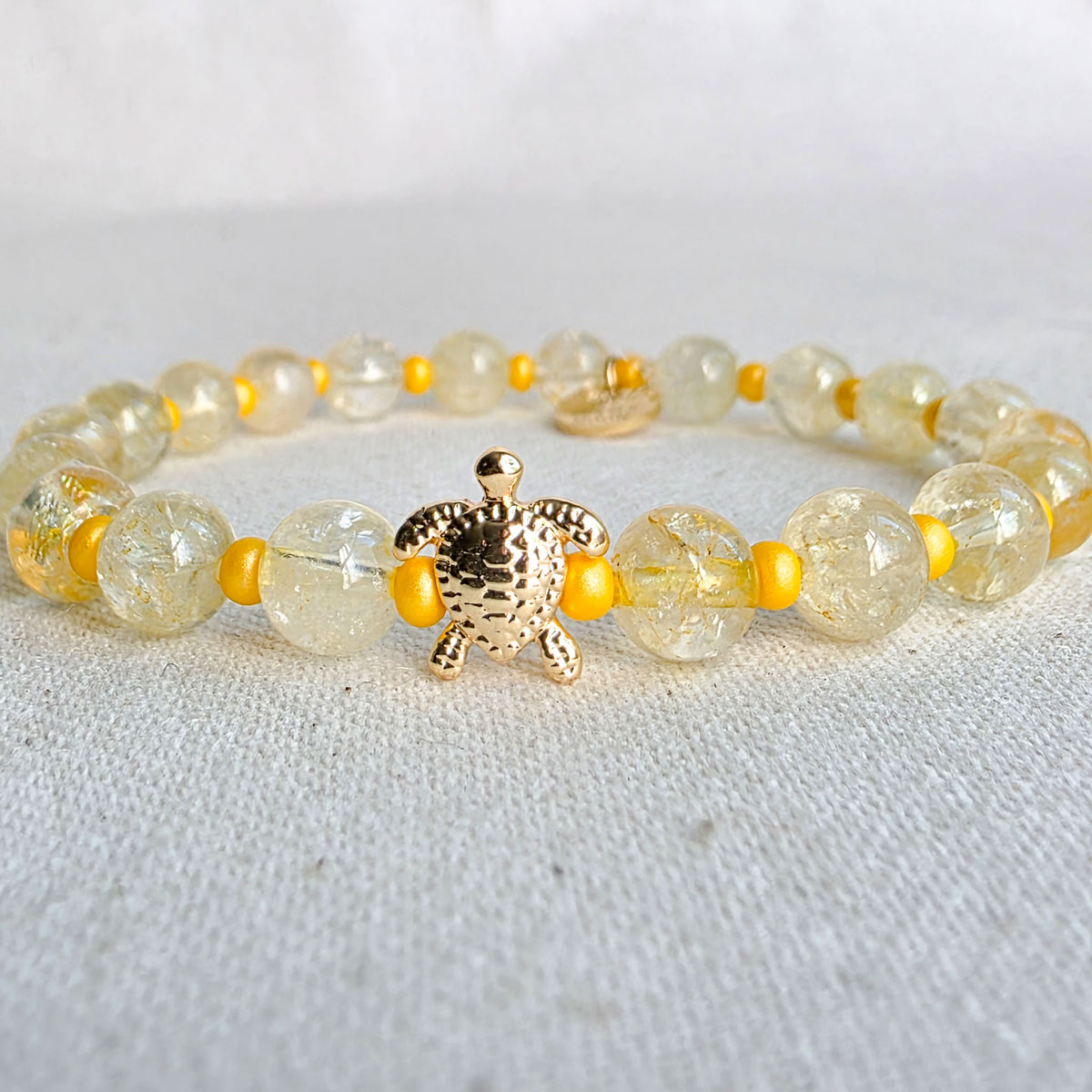 A beaded stretch bracelet featuring round, translucent yellow-green beads with gold foil flakes, small bright yellow spacer beads, and a textured gold turtle charm at the center, displayed on a light fabric background.