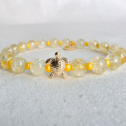A beaded stretch bracelet featuring round, translucent yellow-green beads with gold foil flakes, small bright yellow spacer beads, and a textured gold turtle charm at the center, displayed on a light fabric background.