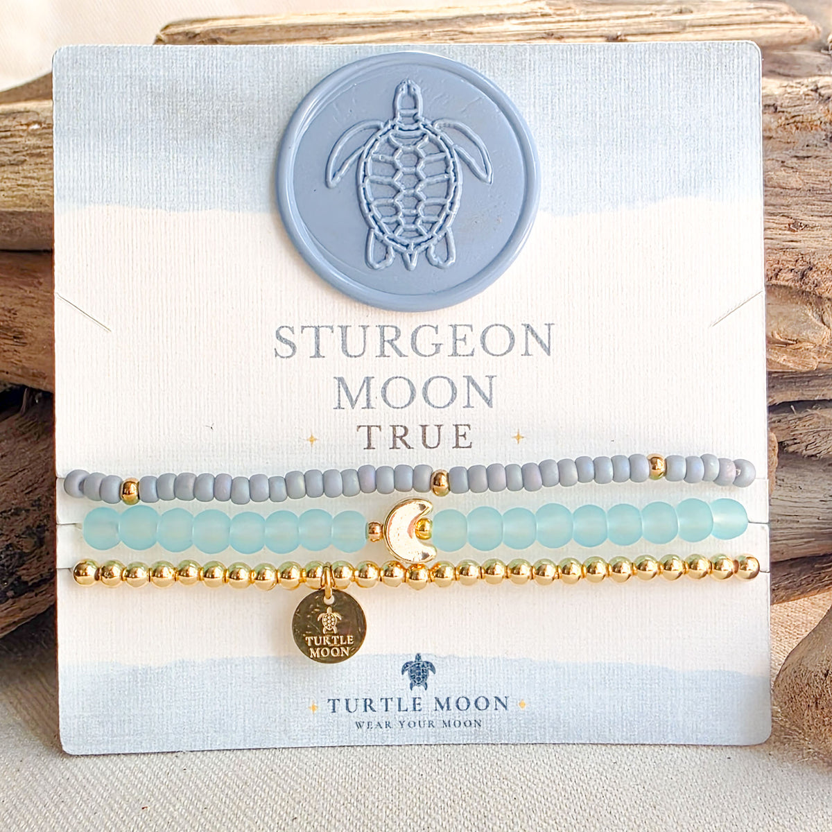 Three beaded stretch bracelets displayed on a branded Turtle Moon card. The bracelets include gray beads, light aqua beads, and gold beads, with a small gold moon charm and a round gold Turtle Moon tag.