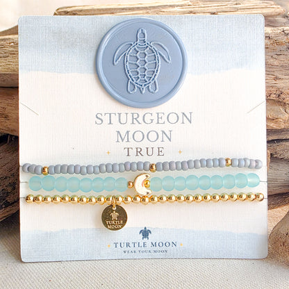 Three beaded stretch bracelets displayed on a branded Turtle Moon card. The bracelets include gray beads, light aqua beads, and gold beads, with a small gold moon charm and a round gold Turtle Moon tag.