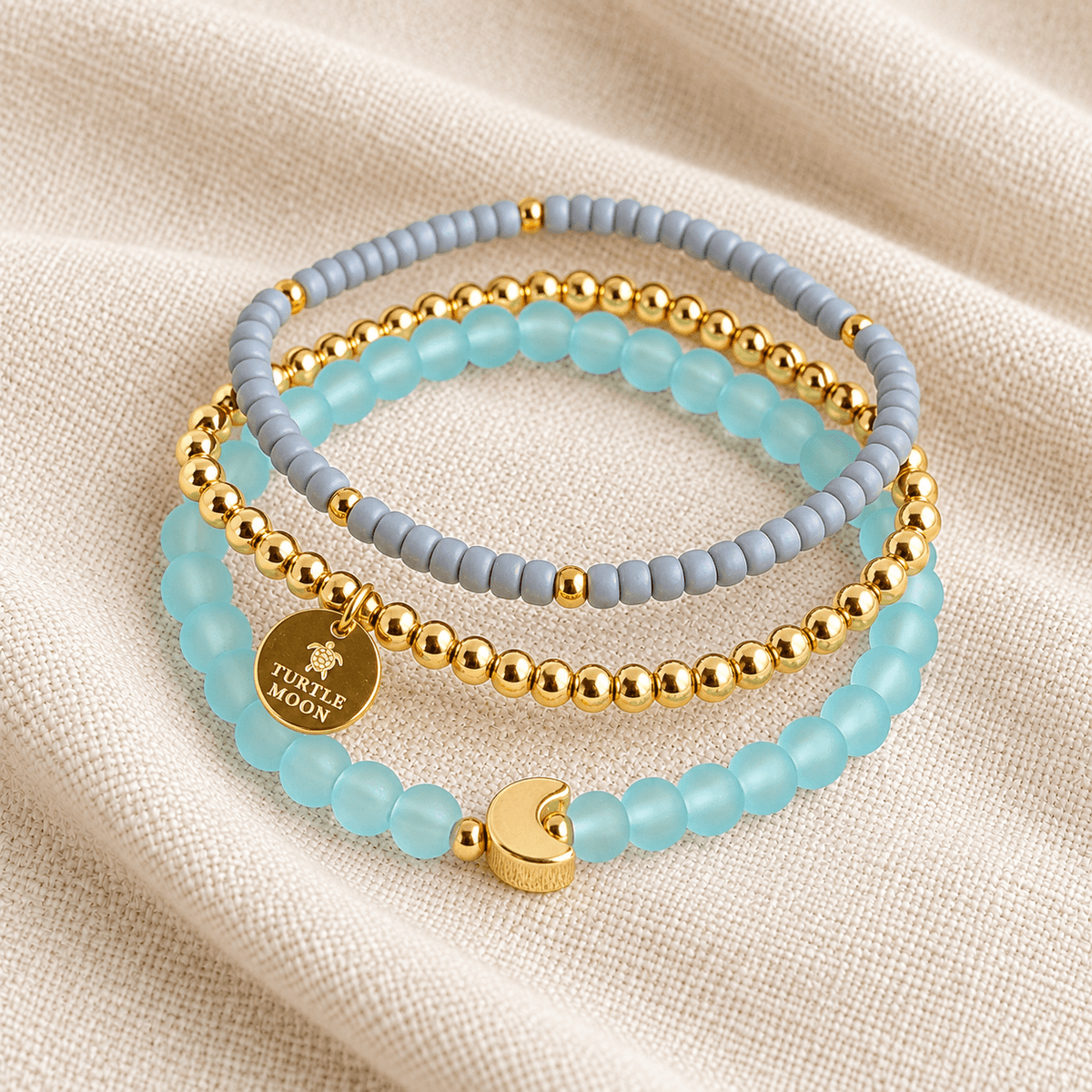 Three beaded stretch bracelets stacked on a textured beige fabric background. The bracelets feature light blue translucent beads, matte gray beads, and shiny gold metallic beads, with one bracelet displaying a round gold charm engraved with 'TURTLE MOON' and a crescent moon accent.