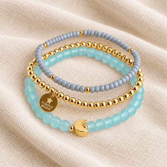 Three beaded stretch bracelets stacked on a textured beige fabric background. The bracelets feature light blue translucent beads, matte gray beads, and shiny gold metallic beads, with one bracelet displaying a round gold charm engraved with 'TURTLE MOON' and a crescent moon accent.