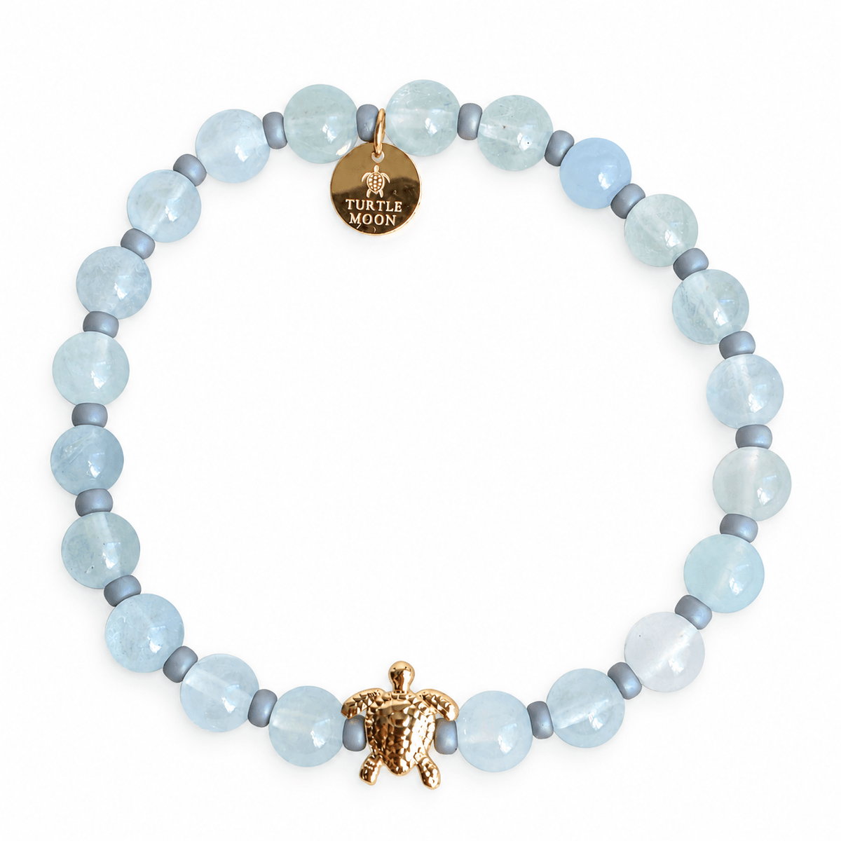 A beaded stretch bracelet featuring light blue aquamarine gemstone beads, small blue gray spacer beads, a textured gold turtle charm, and a round gold disc charm engraved with 'TURTLE MOON'.