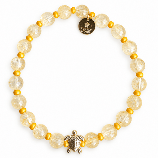 A beaded stretch bracelet featuring citrine quartz crystal beads, small yellow  spacer beads, a textured gold turtle charm, and a round gold disc charm engraved with 'TURTLE MOON'.